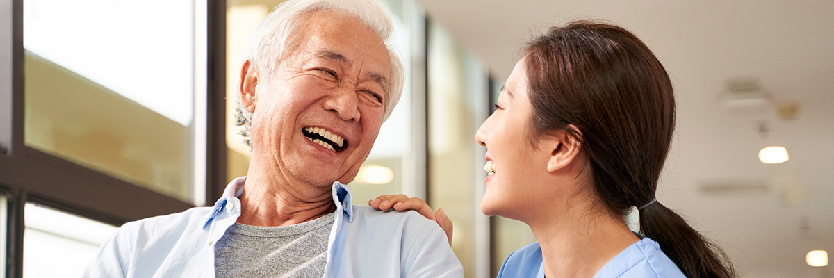 young friendly asian female caregiver talking chatting to happy senior man in hallway of nursing home