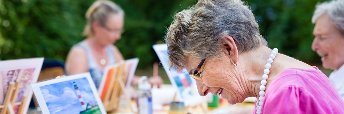 Side view of a happy senior woman smiling while drawing as a recreational activity or therapy outdoors together with the group of retired women.